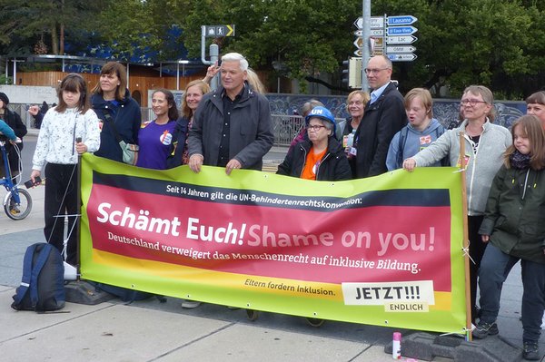 Rund 15 Personen stehen hinter einem Transparent mit der Aufschrift „Schämt Euch! Shame on you! Deutschland verweigert das Menschenrecht auf inklusive Bildung. Eltern fordern Inklusion. Jetzt! Endlich!!" 