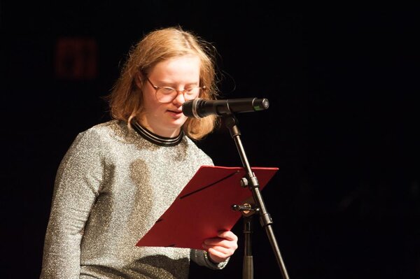 Junge Frau mit Down-Syndrom spricht in ein Mikrofon, sie liest einen Text ab, sie ist durch einen Scheinwerfer erleuchtet