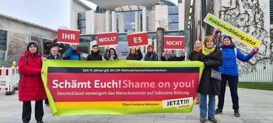 Rund 11 Erwachsene stehen hinter einem Transparent mit der Aufschrift: Seit 14 Jahren gilt die UN-Behindertenrechtskonvention: Schämt Euch! Shame on you! Deutschland verweigert das Menschenrecht auf inklusive Bildung. Eltern fordern Inklusion. Jetzt! Endlich!!!