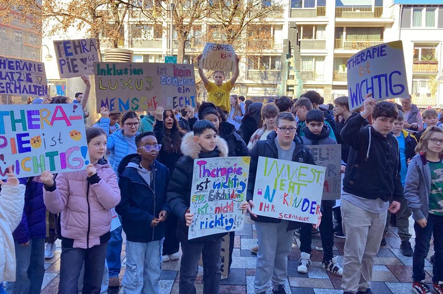 Viele Jugendlcihe setehn dicht beieinander und halten selbst gemalte Schilder mit folgenden Aufschriften vor sich: Therapie ist wichtig; Inklusion ist kein Luxus, sondern Menschenrecht; Investiert in Kinder, Ihr Schwindler!