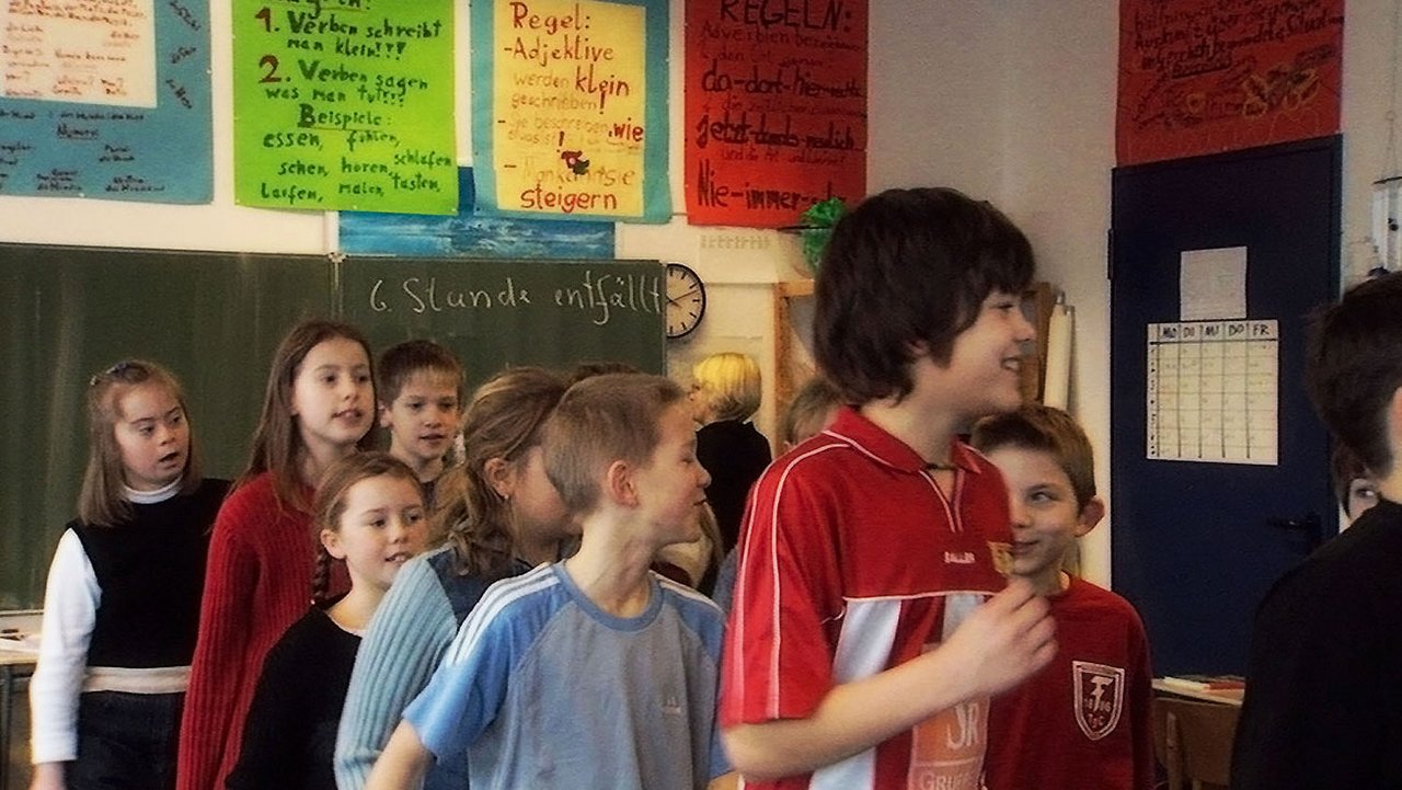 Kinder im Durcheinander in einer Klasse stehend, im Hintergrund eine Schultafel und bunte Plakate an den Wänden.