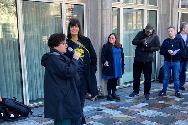 Eine Person spricht mit einem Mikrofon vor einem Gebäude zu einer kleinen Gruppe, während mehrere Erwachsene daneben stehen und zuhören.