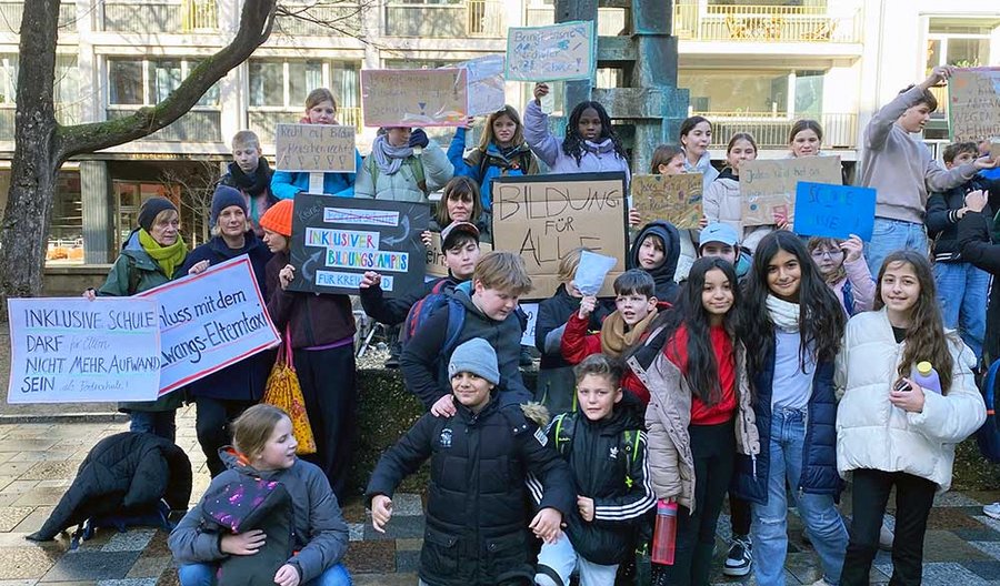 Etwa 40 Jugendliche und Erwachsene stehen zusammen und halten Schilder in der Hand. Auf den Schildern steht: Bildung für alle, Inklusiver Bildungscampus für Kreuzfeld, Schluss mit dem Zwangs-Elterntaxi, Inklusive Schule darf für Eltern nicht mehr Aufwand sein als Förderschule! Recht auf Bildung = Menschenrecht, Bringt unserer Mitschüler in die Schule!