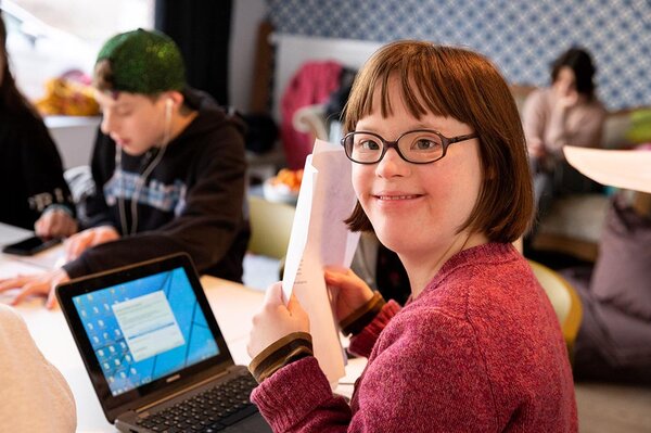 Mädchen mit Down-Syndrom sitzt am Tisch mit Blättern in der Hand vor sich einen Laptop, im Hintergrund weitere Jugendliche am Tisch beim Schreiben.