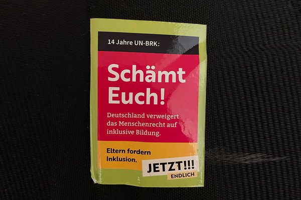Ein etwas zerknitterter Aufkleber klebt auf dunklen Hintergrund. Auf Flächen in Schwarz Rot und Gelb steht: 14 Jahren UN-BRK: Schämt Euch! Shame on you! Deutschland verweigert das Menschenrecht auf inklusive Bildung. Eltern fordern Inklusion. Jetzt! Endlich!!!“