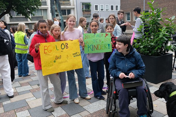 Eine Gruppe junger Demonstrierender posiert mit selbst gemalten Plakaten. Darauf stehen „Egal ob Gesamtschule oder Hauptschule“ und „Nicht auf Kosten der Kinder!“. Rechts sitzt eine lachende junge Person im Rollstuhl.