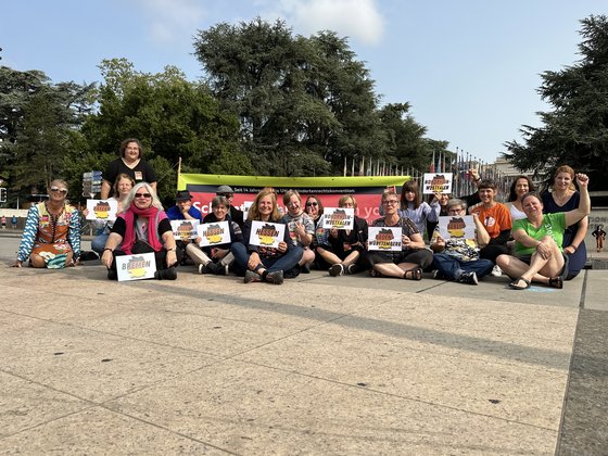 Eine Gruppe von Menschen sitzt auf dem Platz vor dem Palais des Nations in Genf, sie halten Schilder mit den Namen der Bundesländer in der Hand, aus denen sie kommen: NRW, Hessen, Bremen, Berlin, Baden-Württemberg, Bayern