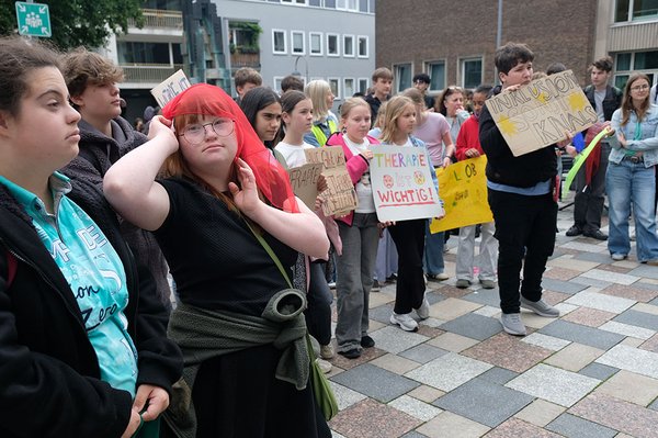 Jugendliche mit und ohne Behinderungen versammeln sich mit bunten Protestschildern für Inklusion und Therapieangebote. Im Vordergrund trägt eine junge Frau mit rotem Haar ein rotes Tuch über dem Kopf.