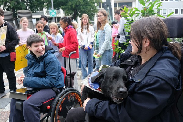 Zwei junge Rollstuhlnutzende lächeln einander an. Auf dem Schoß der Person rechts liegt ein schwarzer Assistenzhund; im Hintergrund stehen Jugendliche mit Demonstrationsplakaten.