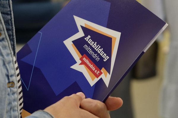 Man sieht die Tagungsmappe mit dem Logo des Projekts „Ausbildung mittendrin“. Sie wird von einer Teilnehmer:in in der Hand gehalten.