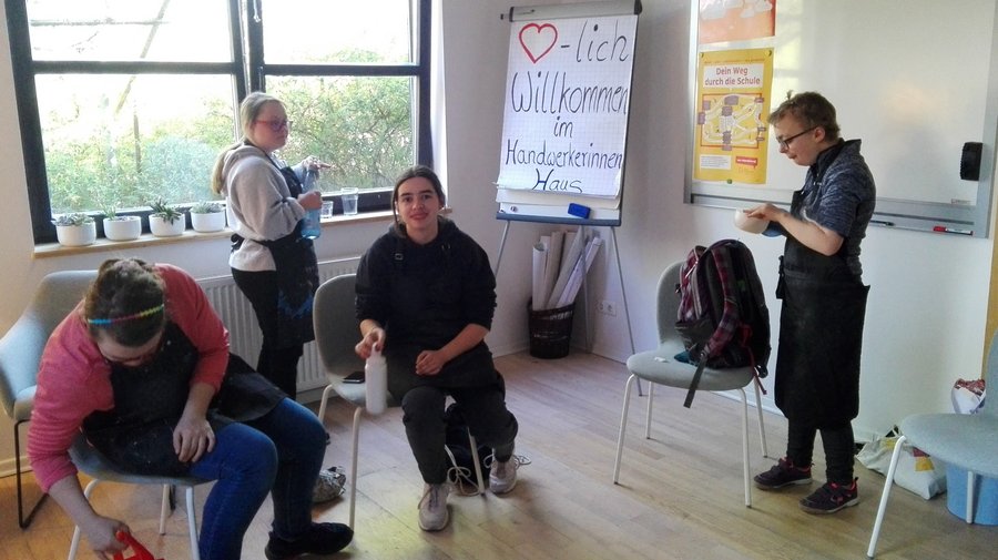 Schnupperkurs im Handwerkerinnenhaus Das Bild zeigt einen Raum mit vier Personen, die verschiedenen Aktivitäten nachgehen. Der Raum verfügt über einen Holzboden und ist mit Stühlen und einem Whiteboard ausgestattet. Auf dem Whiteboard steht Herzlich Willkommen.