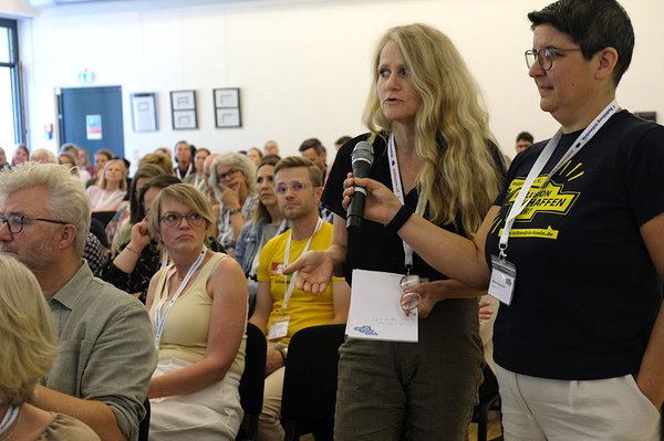Man sieht das Publikum. Eine Teilnehmerin steht am Rand und spricht ins Mikrofon. Eine Frau vom Team des mittendrin e.V. hält ihr das Mikro hin.