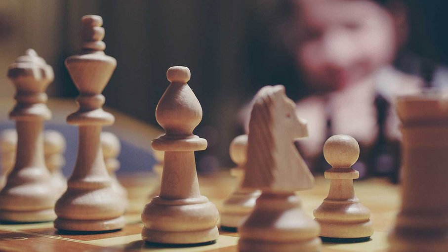 Bick_Schach_web Weiße Figuren auf einem Schachbrett, im Hintergrund ist verschwommen ein Mensch zu erkennen.