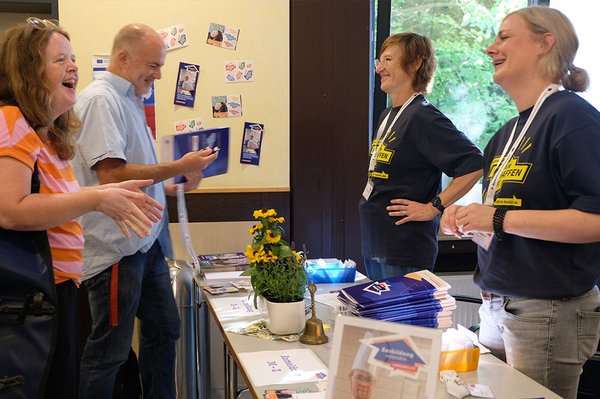 Man sieht vier Personen, zwei Kongressteilnehmer:innen und zwei Mitglieder des mittendrin e.V. Zwischen Ihnen der Anmeldetisch mit Mappen und Namensschildern. Im Hintergrund an der Wand Projekt-Flyer, Logo-Postkarten und das Förderplakat des Europäischen Sozialfonds. Die Personen unterhalten sich und lachen.