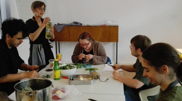 Das Bild zeigt eine Gruppe von fünf Personen, die um einen rechteckigen Tisch sitzen und stehen. Der Tisch ist mittig im Raum positioniert, und die Personen sind mit der Zubereitung der Speisen beschäftigt.