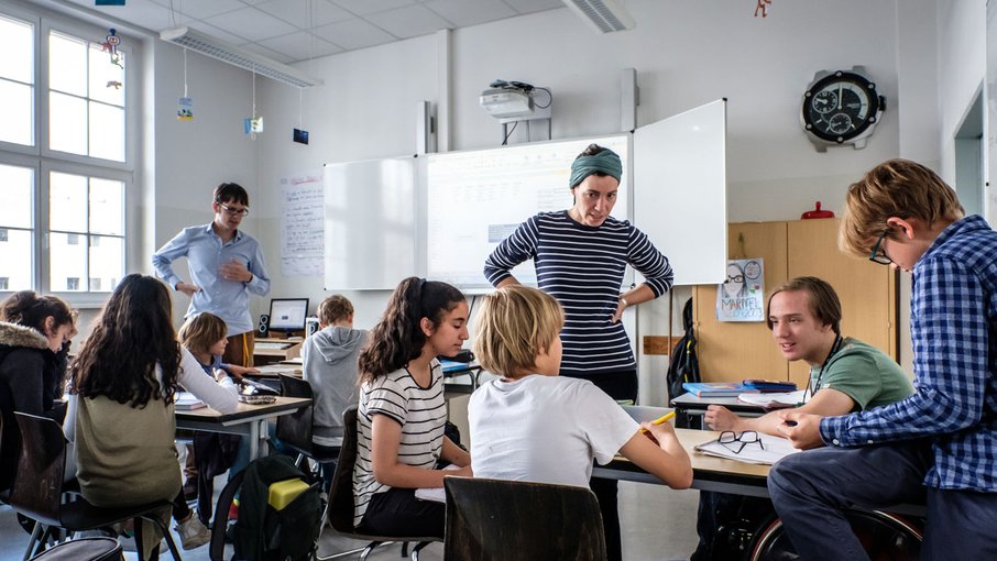 Klassenzimmer mit Schüler:innen Etwa 10 Schüler in einem Klassenzimmer an Tischen sitzend. Zwei Lehrer erklären ihnen etwas. Im Hintergrund ein großes Whiteboard.