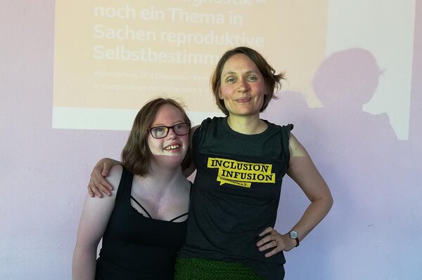 Zwei Frauen, eine davon mit Down-Syndrom, die Arme umeinander gelegt, vor einer Wand mit darauf projizierter Präsentation. 