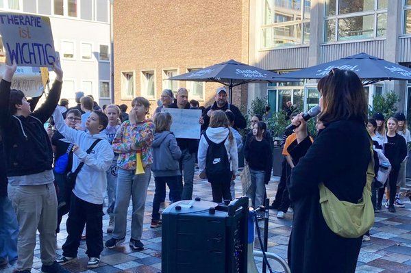 Eine Frau spricht mit einem Mikrofon zu einer Gruppe von Kindern und Erwachsenen bei einer Kundgebung im Freien. Mehrere Teilnehmende halten Plakate hoch, während die Zuhörenden sich um die Rednerin versammeln.