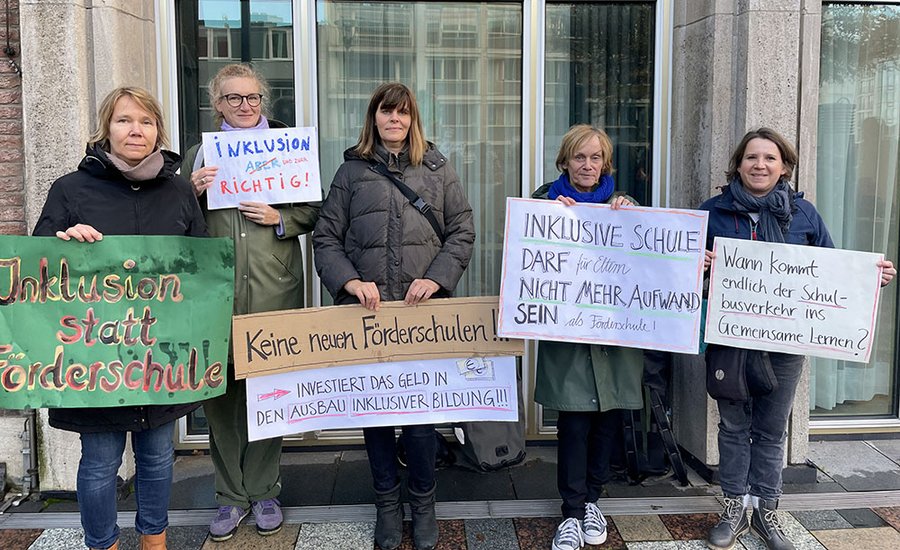 Fünf Personen stehen nebeneinander vor einem Gebäude. Sie halten Schilder vor sich mit folgenden Aufschriften: Inklusion statt Förderschulen, Inklusion und zwar richtig, Keine Neuen Förderschulen! Investiert das Geld in den Ausbau inklusiver Bildung!, Inklusive Schule darf für Eltern nicht mehr Aufwand sein als Förderschule. Wann kommt endlich der Schulbusverkehr ins Gemeinsame Lernern?