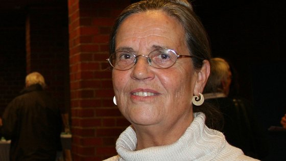 Eine Frau mittleren Alters trägt eine Brille mit dünnem Gestell und einen weißen Rollkragenpullover.