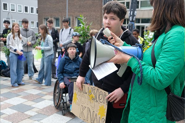 Ein Jugendlicher spricht bei der Demonstration in ein Mikrofon und hält ein Schild mit der Aufschrift „Inklusion – sonst knallt’s“. Eine Frau hält ein Megafon; im Hintergrund stehen viele Jugendliche, eine der Personen sitzt sitzt ein im Rollstuhl.
