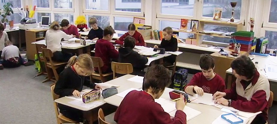 Schüler im Klassenzimmer Etwa 10 Kinder im einem Klassenzimmer an Gruppentischen sitzend. Sie sind jeweils über ein Blatt Papier gebeugt.