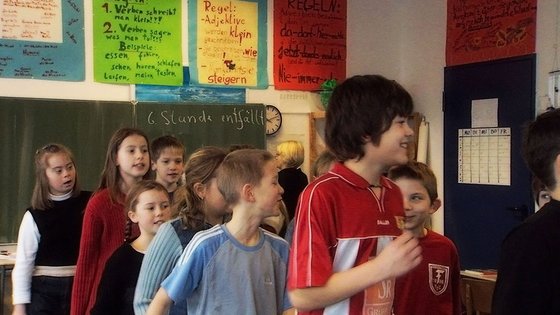 Kinder im Durcheinander in einer Klasse stehend, im Hintergrund eine Schultafel und bunte Plakate an den Wänden.