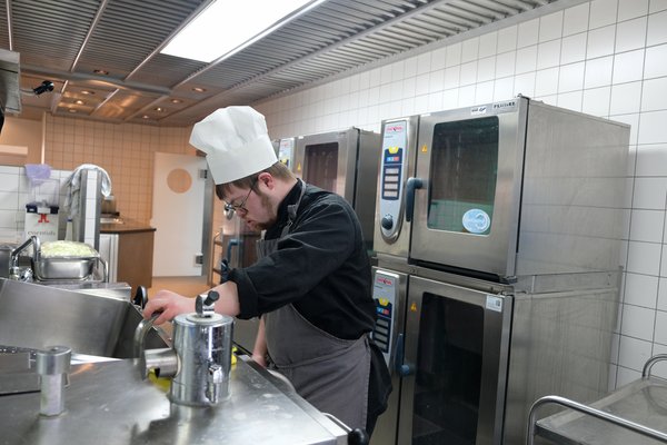 Das Bild zeigt eine professionelle Küche mit einem arbeitenden Koch. Der Koch trägt eine weiße Kochmütze, eine schwarze Kochjacke und eine graue Schürze.