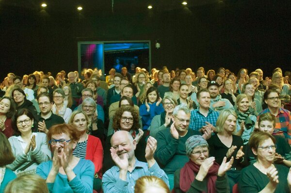 Publikum in voll besetztem, beleuchtetem Theatersaal beim Applaus, lachende Gesichter.