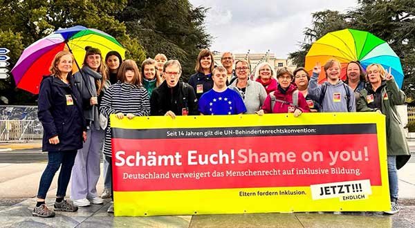Rund 20 Erwachsene und Jugendliche stehen mit bunten Regenschirmen hinter einem Transparent mit der Aufschrift: Seit 14 Jahren gilt die UN-Behindertenrechtskonvention: Schämt Euch! Shame on you! Deutschland verweigert das Menschenrecht auf inklusive Bildung. Eltern fordern Inklusion. Jetzt! Endlich!!!