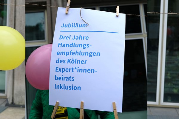 Zwischen bunten Luftballons hängt ein Plakat mit der Aufschrift: „Jubiläum – Drei Jahre Handlungsempfehlungen des Kölner Expert*innenbeirats Inklusion“