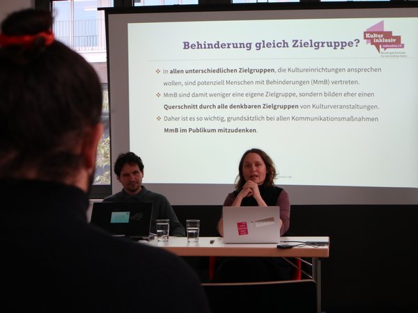 Adriani Botez und Tina Sander an Tisch hinter aufgeplappten Laptops. Hinter ihnen die Projektion einer Präsentationsfolie mit Headline: Behinderung gleich Zielgruppe?