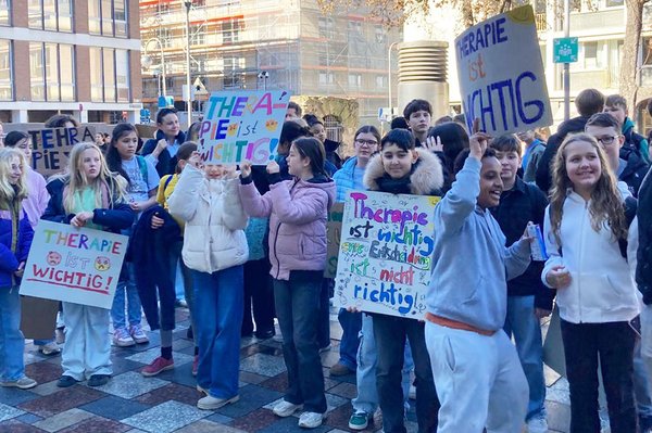 Kinder und Jugendliche stehen dicht beieinander bei einer Demonstration und zeigen bunte Plakate mit Forderungen wie „Therapie ist wichtig“ in die Kamera.