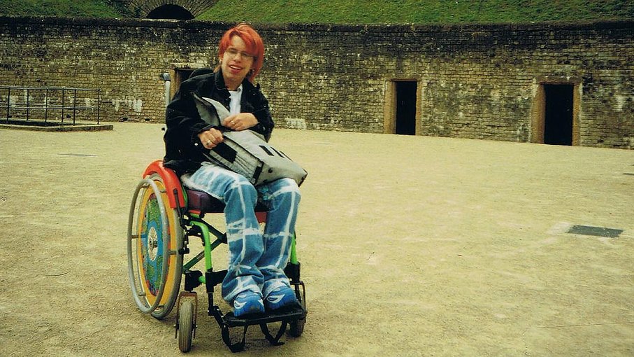 Junge Person im Rollstuhl Eine junge Person mit roten Haaren im Rollstuhl. Sie trägt ein dunkles Oberteil und eine Jeanshose und hält eine Tasche auf dem Schoß.