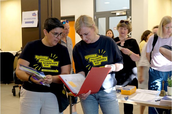 Im Vordergrund stehen zwei Frauen vom Team des mittendrin e.V. und schauen etwas gemeinsam in einem Ordner nach. Sie tragen dunkelblaue T-Shirts mit dem leuchtend gelben Logo „mittendrin e.V.. Inklusion schaffen wir!“. Im Hintergrund sieht man Tagungs-Teilnehmer am Anmeldetisch.