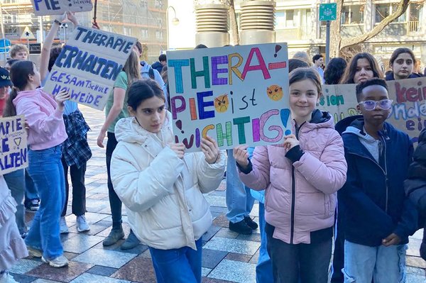 Kinder und Jugendliche laufen gemeinsam bei einer Demonstration und halten bunte Plakate mit Aufschriften wie „Therapie ist wichtig“ in die Kamera.