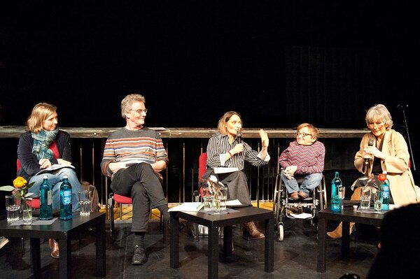 Fünf Teilnehmer:innen einer Diskussion sitzen auf einer beleuchteten Bühne, vier Frauen und ein Mann, eine Frau ist Rollstuhlfahrerin