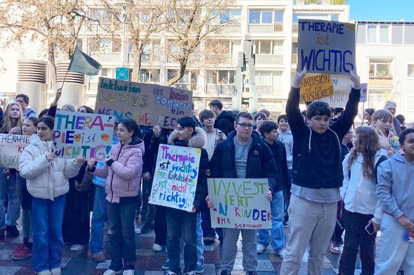 Eine große Gruppe von Kindern und Jugendlichen demonstriert auf einer Straße und hält Schilder mit Aufschriften wie „Therapie ist wichtig“ und „Inklusion ist kein Luxus“ hoch.