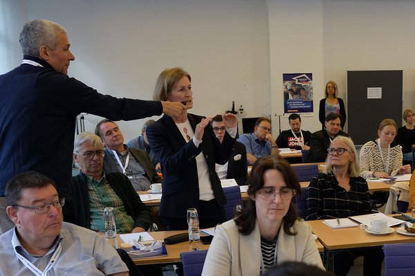 Blick in den Saal, Publikum von vorne, zwei Personen stehen, der stehende Mann links hält der stehenden Frau in der Mitte das Mikrofon hin 
