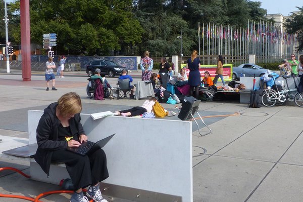 Eine Person sitzt auf einer Bank und schreibt an einem Laptop. Im Hintergrund ist das Protestcamp mit Transparent und Menschen zu sehen.