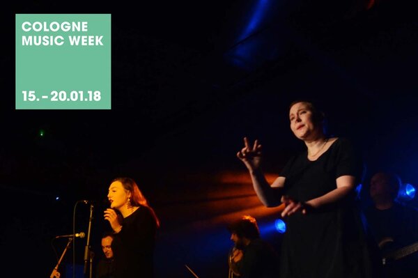 Sängerin auf beleuchteter Bühne, daneben eine Gebärdensprachdolmetscherin, oben in der Ecke eine grüne Farbfläche mit Text: Cologne Music Week, 15.-20.01.18