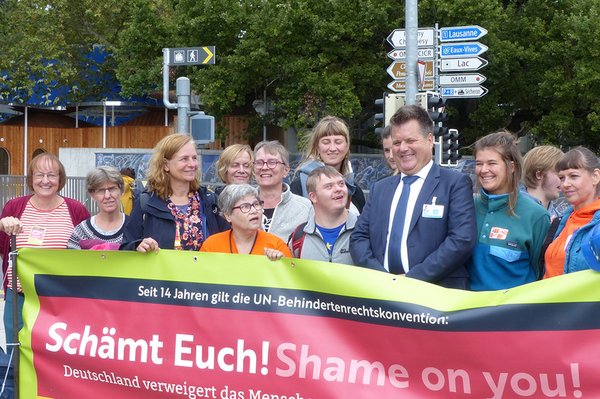 Rund 15 Personen stehen lachend hinter dem Transparent mit der Aufschrift „Schämt Euch! Deutschland verweigert das Recht auf inklusive Bildung.“