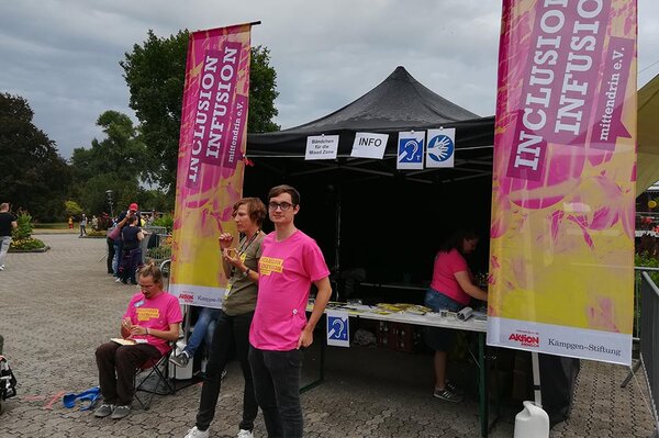 Ein schwarzer Pavillon auf einem Open-Air-Konzert-Gelände, davor einige Personen. Rechts und links vom Pavillon große Fahnen mit aufgedrucktem Logo:Inclusion Infusion. mittendrin e.V. Am Pavilliondach Schilder mit Icons für Gebärdensprache und Induktionstechnik.