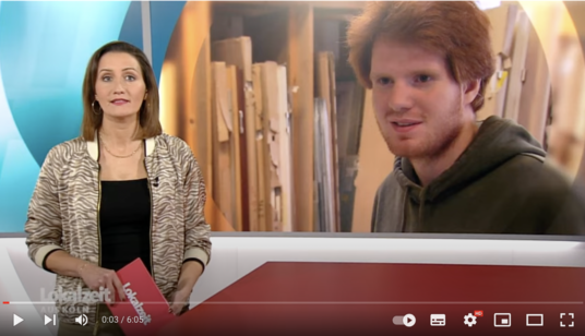 Screenshot des Youtube-Videos: Links im Bild die Moderatorin, dahinter eingebkendet Dominik, ein junger Mann mit roten Haaren, er steht vor einem Regal mit Holz und lächelt.