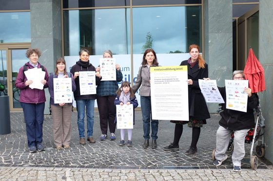 Ein Gruppe von sieben Erwachsenen und einem Kind steht vor einem Regierungsgebäude. Jede Person hält einen Teil des offenen Briefe in der Hand. 