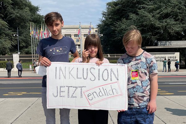 Drei Jugendliche stehen nebeneinander und halten ein Plakat mit der Aufschrift: Inklusion Jetzt Endlich! Zwei der Jugendlichen haben Down-Syndrom.