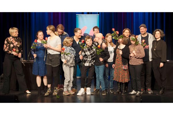 Gruppe von Menschen auf erleuchteter Bühne beim Schlussapplaus, alle haben eine Blume in der Hand, fröhliche Gesichter