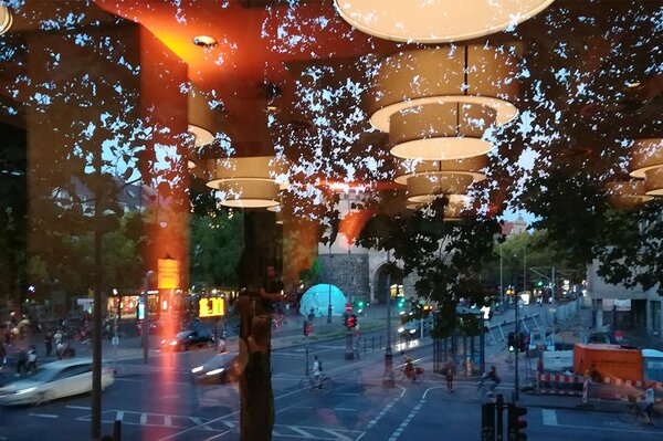 Durch eine Scheibe mit reflektierenden Lampen der Blick auf eine städtische große Kreuzung mit Platz, darauf eine große aufgeblasene Hülle, wie eine Blase, die von innen blau leuchtet.