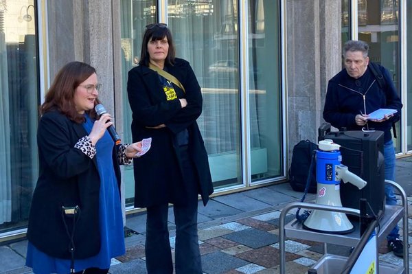 Eine Frau spricht bei einer Kundgebung mit einem Mikrofon vor einem Gebäude, während zwei weitere Personen neben ihr stehen und ein Megafon sowie Technik bereitstehen.