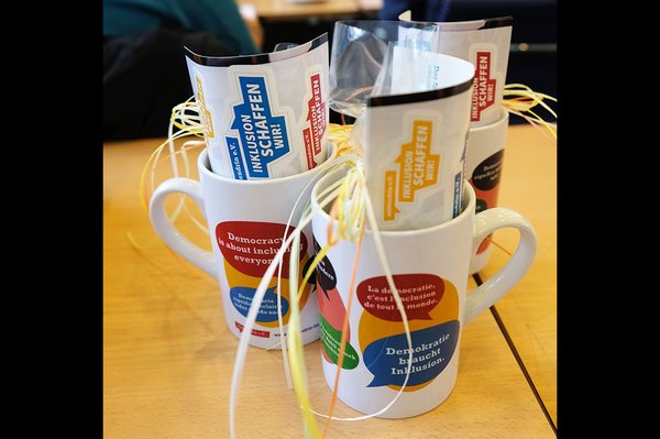 3 Tassen als Präsent hergerichtet. Auf den den Tassen steht der Print Inklusion braucht Demokratie in verschiedenen Sprachen.